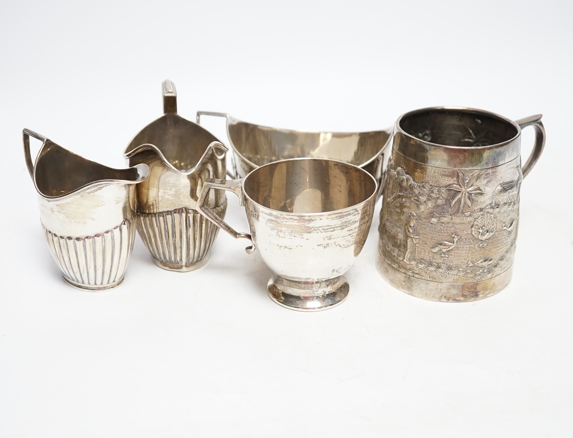 Two early 20th century demi-fluted silver cream jugs and a similar sugar bowl, an Irish silver tea cup and an Indian? white metal mug, gross weight 16.7oz. Fair condition.